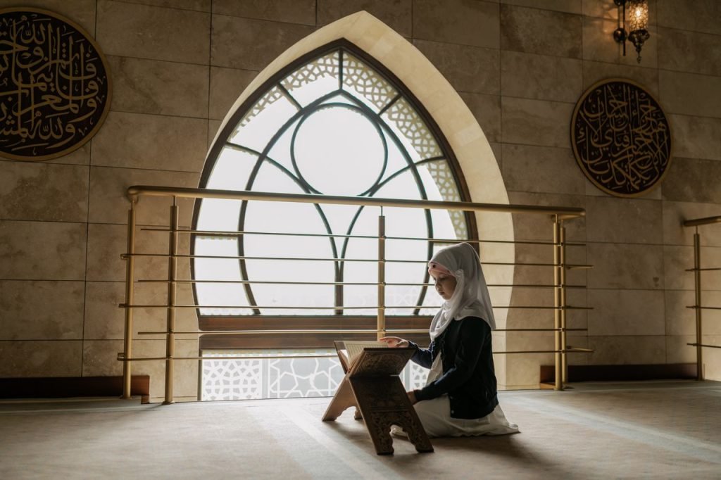 A woman in hijab prays and reads the Quran in a beautifully lit mosque interior.