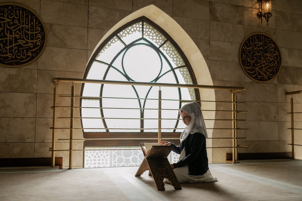 A woman in hijab prays and reads the Quran in a beautifully lit mosque interior.